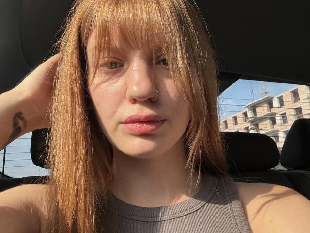 ElsaBj with dirty blonde hair, nose ring, and small tattoo on right wrist, wearing a gray tank top, sitting in a car, sunlight on her face, looking at camera.