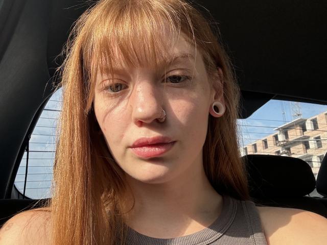 ElsaBj has red hair, a nose ring, and white ear gauges. She's wearing a gray tank top, with light freckles on her face, and pinkish lips.
