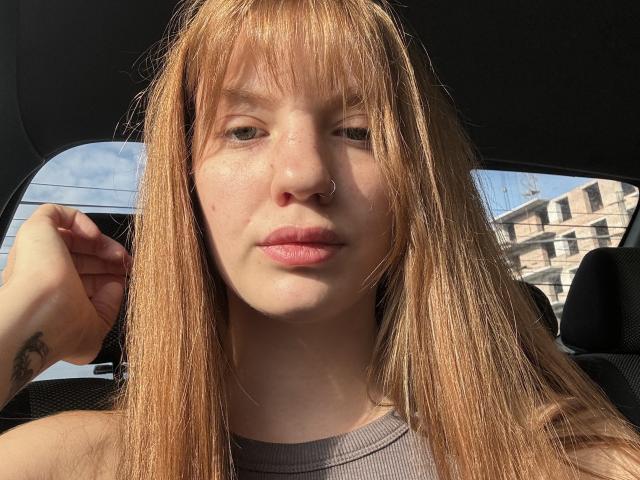ElsaBj with long, straight, blonde hair, nose ring, and tattoo on her left wrist, inside a car, sunlight on her face, wearing a gray top, looking at the camera.