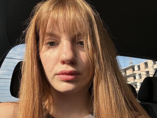 ElsaBj has straight, dirty blonde hair with bangs, a nose ring, and pale skin. She's sitting in a car, looking serious with pink lips. Sunlight hits her face.