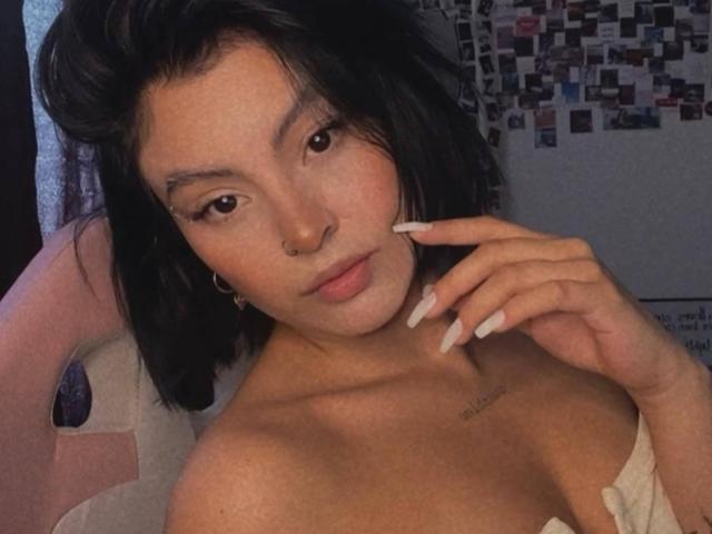 EimyBelle with black shoulder-length hair, nose ring, and long white nails, wearing off-shoulder top, touching chin, tattoo on right collarbone, looking at camera.