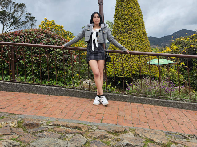 NicoColeman stands with arms on a railing, wearing a gray jacket, white shirt, black shorts, and white sneakers. She's got a cute ass and a relaxed, confident look.