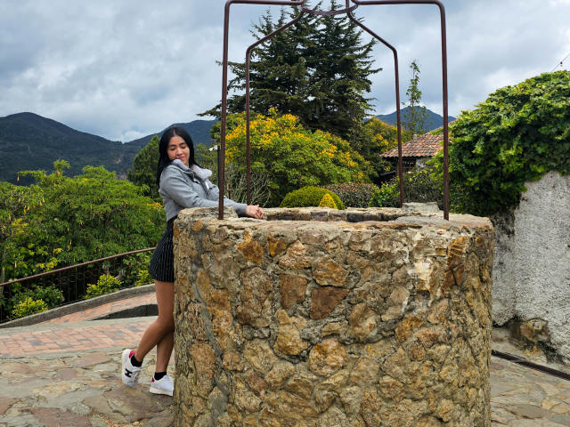 NicoColeman with black hair, grey hoodie, and black shorts, leaning on a stone water pump, wearing white sneakers, outdoors, cloudy sky, mountains in background, holding a playful expression.