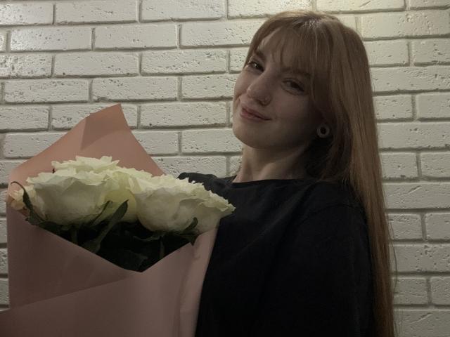ElsaBj with long red hair, black shirt, holding pink-wrapped bouquet of white roses, smiling slightly, ear gauge visible, dim lighting, white brick wall in background.