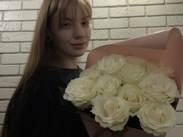 ElsaBj with pale skin and long red hair, holding a bouquet of white roses, wearing a black shirt, and a subtle smile, with ear gauges visible.