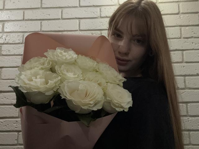 ElsaBj with long red hair, holding a pink bouquet of white roses, wearing a black shirt, nose ring, and subtle makeup, looking slightly to the side.
