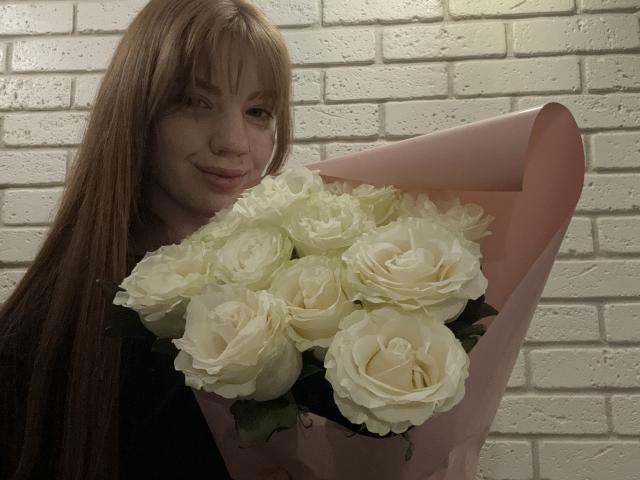 ElsaBj with long brown hair, holding a pink bouquet of white roses, smiling subtly. Her tits are slightly visible under her black top. Her face is lightly flushed.