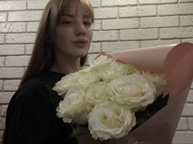 ElsaBj with red hair, black shirt, holding white roses in pink paper, looking at camera, subtle smile, white brick wall background, small black ear gauges.