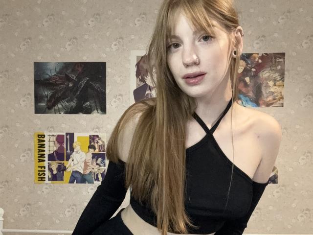 ElsaBj with long brown hair, nose ring, black off-shoulder top, and ear gauges, showing a cute face and small tits, standing in a room with posters.