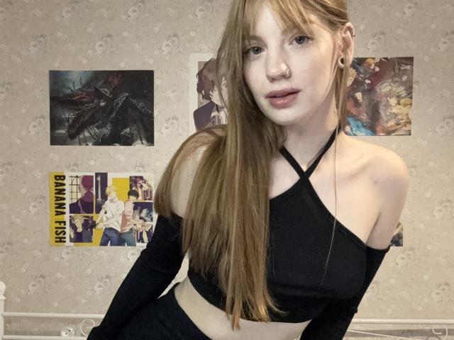 ElsaBj with long brown hair, black off-shoulder top, and black pants, showing her bare shoulders and a small nose ring, in a room with posters on a beige wall.