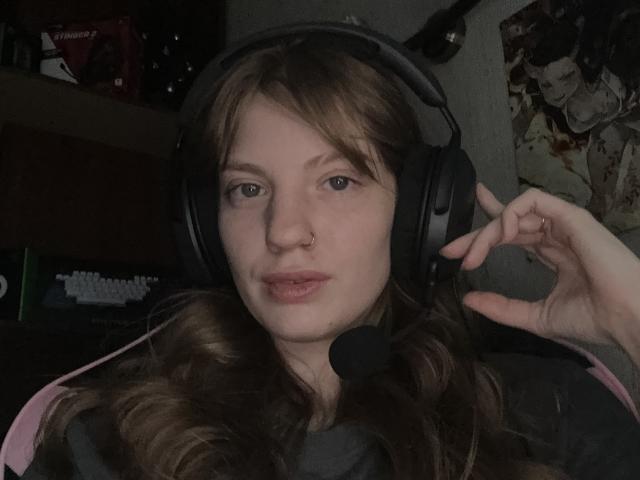 ElsaBj with wavy brown hair, wearing black headphones, a nose ring, and a headset, making a 'shushing' gesture with her right hand, in a dimly lit room.