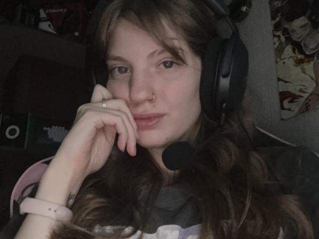 ElsaBj with wavy brown hair, wearing black headphones, a nose ring, and a pink wristband, resting her chin on her hand, smiling subtly.