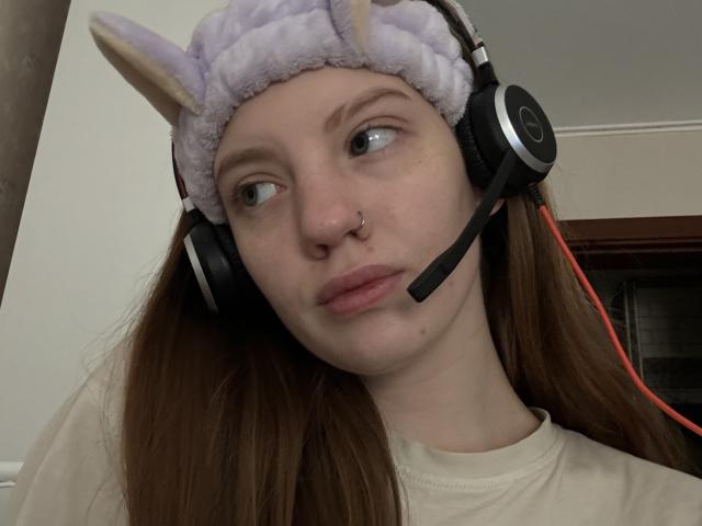 ElsaBj has light skin, brown hair, wearing a purple bunny headband, black headphones, and a white shirt, with a nose ring, looking slightly bored, with a small red headset cord.