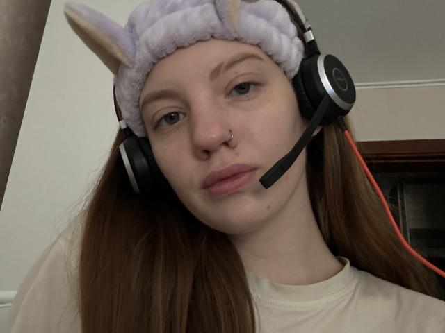 ElsaBj with brown hair, wearing a fluffy purple cat ear headband, black headphones with a mic, and a nose ring, looking serious in a selfie.