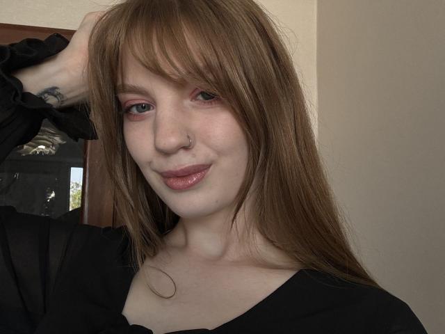 ElsaBj has light brown hair, pink eye makeup, a nose ring, and is wearing a black top, with a slight smile and one arm raised. Her tits are small.