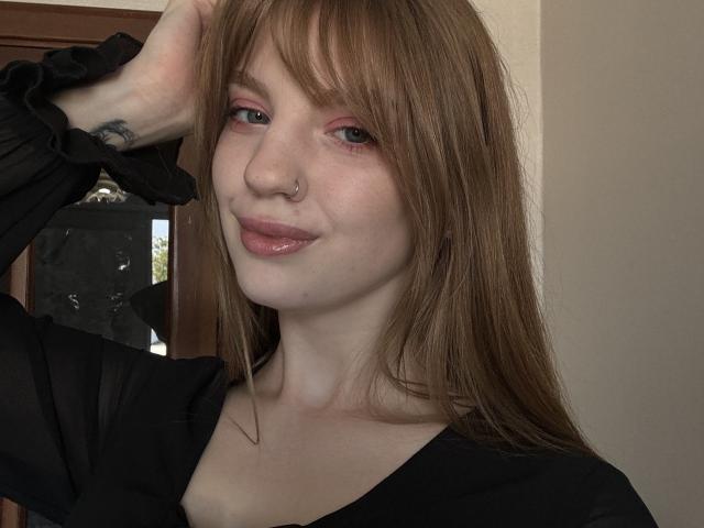 ElsaBj has long red hair, pink eye makeup, a nose ring, and a black top, smiling with glossy pink lips, right arm raised, showing a tattoo on her wrist.