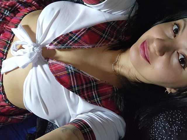 AlySaviny69 with small tits, wearing a red plaid shirt tied at the front, white top, and plaid skirt, lying down, smiling, dark hair, pink lipstick, gold necklace.