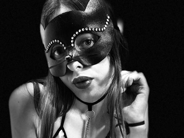 BlueViolette with a black mask adorned with studs, wearing a choker with a heart, and looking at the camera with pouty lips, in a black-and-white photo.