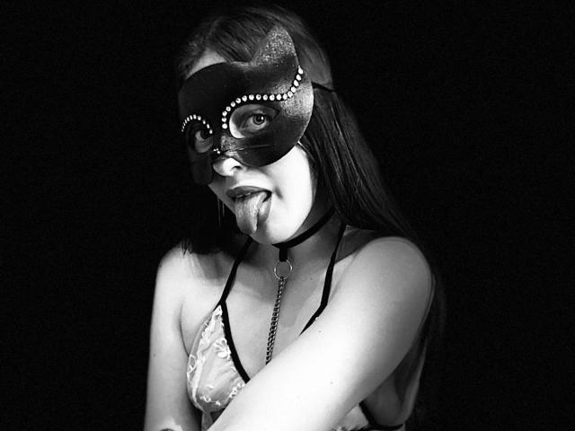 BlueViolette with a mask, tongue out, drooling, wearing a lace bra, black choker, and chain, in black-and-white, looking sexy. Her tits are visible, and she's hot.