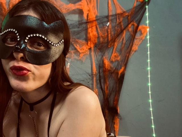 BlueViolette with a black mask, red lipstick, and brown hair, wearing a choker and black halter, has her pouty lips slightly pursed, with orange webbing in the background.