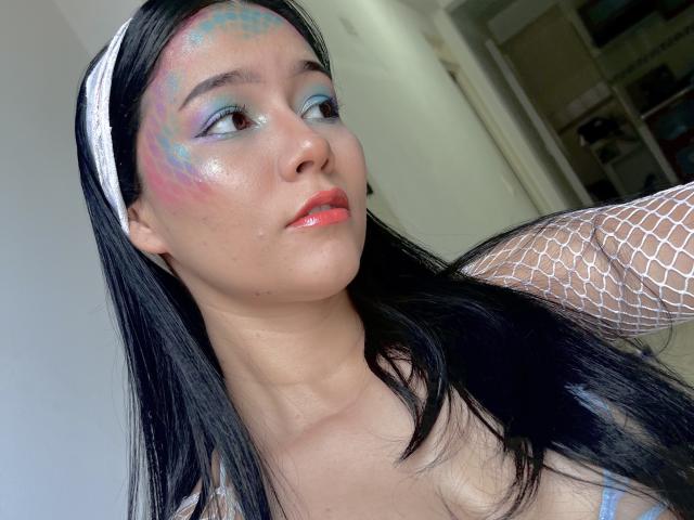 DeisyWest, com lábios rosados e brilhantes, cabelo preto, faixa de cabelo branca, braço com meia-calça de peixe, maquiagem colorida nos olhos e pele luminosa, olhando para o lado com um nariz levemente arrebitado..
