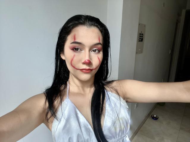 DeisyWest hat langes, schwarzes Haar, rote Make-up-Linien im Gesicht und ein silbernes Halstuch-Top. Ihre Arme sind ausgestreckt und zeigen eine weiße Wand und einen gefliesten Boden..
