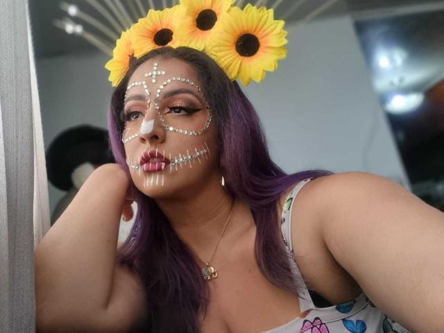 AndreaSharick with purple hair, sunflower headpiece, face painted with cross and rib patterns, wearing a low-cut floral dress, touching her cheek, lipstick, and a necklace. Boobs visible.