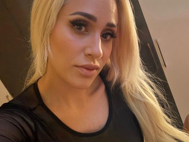 LunaSky-hot has blonde hair, dark eyebrows, long eyelashes, and is wearing a black top. She has a subtle smile and is looking at the camera with a sexy expression.