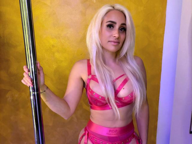 LunaSky, met haar platina blonde haar, roze bandjeslingerie, terwijl ze een paal vasthoudt, haar borsten laat zien, een roze gartersceel draagt en een armband, staand voor een gele muur..