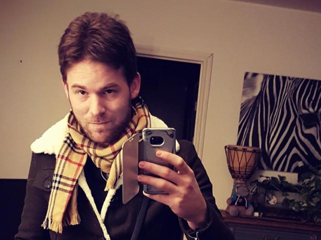 KinkFetish has brown hair, a beard, and a plaid scarf, holding a phone in a mirror selfie, wearing a dark jacket with a white fleece collar.