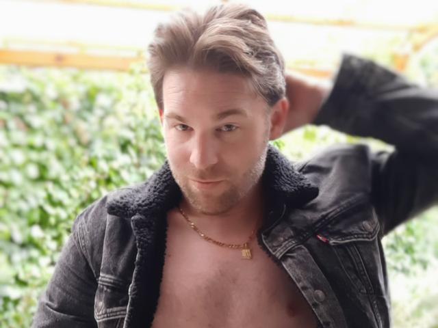 KinkFetish with short brown hair, black denim jacket, chest exposed, gold necklace, hand in hair, slight smile, looking down, casual, sexy, handsome, confident.