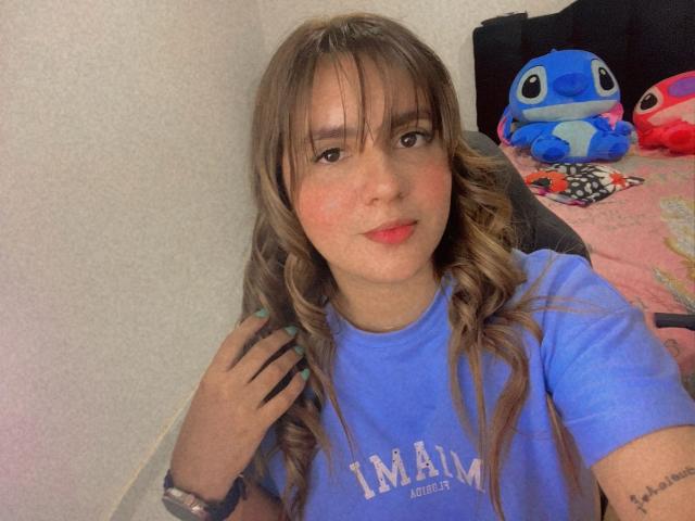 IsaFW with long brown wavy hair, blue shirt, light skin, red lips, small tits, wearing a watch, holding phone, blue-eyed, sitting with blue and red stuffed animals.
