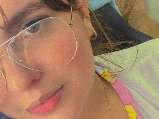 IsaFW's face with pink lipstick, wearing clear glasses and gold hoop earrings, has light brown skin and brown hair, showing a subtle smile. Visible pink and yellow shirt.