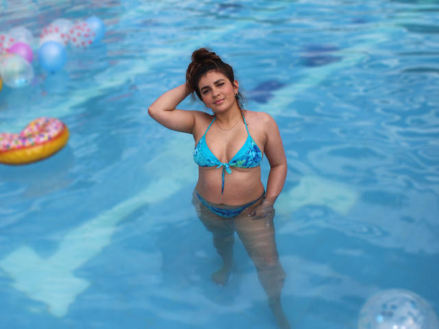 IsaFW in a blue floral bikini, standing in a pool, hand in hair, looking up, water covering her lower body, with a cute butt and small tits.