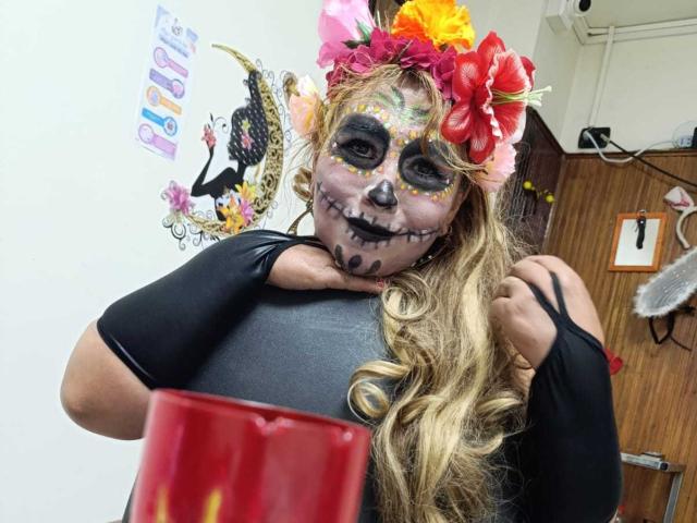 Kazsandra with colorful skull makeup, flowers in her blonde hair, black top, and curly hair, holding red cup, in a room with a wooden door.