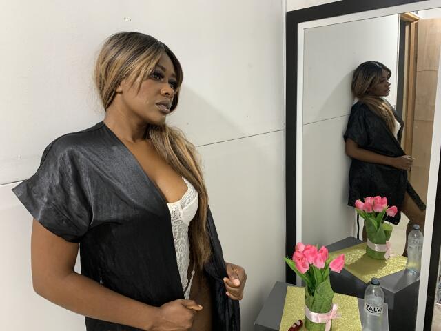 AstridDaviz, com pele morena e cabelos longos loiros, veste um roupão de cetim preto, expondo seu sutiã de renda branca e mostrando seus seios. O reflexo no espelho mostra suas costas..