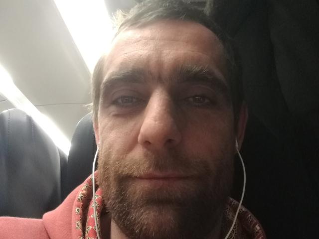 TerzoHot with a slight smile, brown beard, white earbuds, and a red shirt with a pattern, sitting in a dimly lit airplane seat.