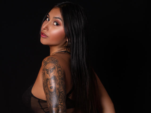 SarahOriz with long black hair, brown skin, and red lipstick, looking over her shoulder, showing a tattooed arm, wearing a sheer black top, against a black background.