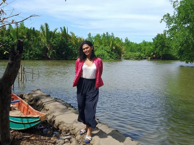 ErikaTS with black hair, medium tits, wearing a pink jacket, white top, and navy pants, standing on a rocky riverbank, green boat in the left foreground, sunny day.
