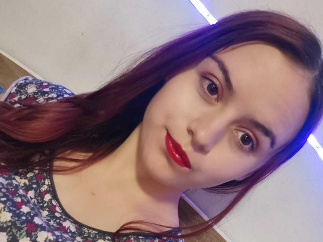 TeenyCouple has fair skin, red lipstick, and brown eyes. She's wearing a floral shirt, with long reddish-brown hair and a slight smile. Her tits are not visible.