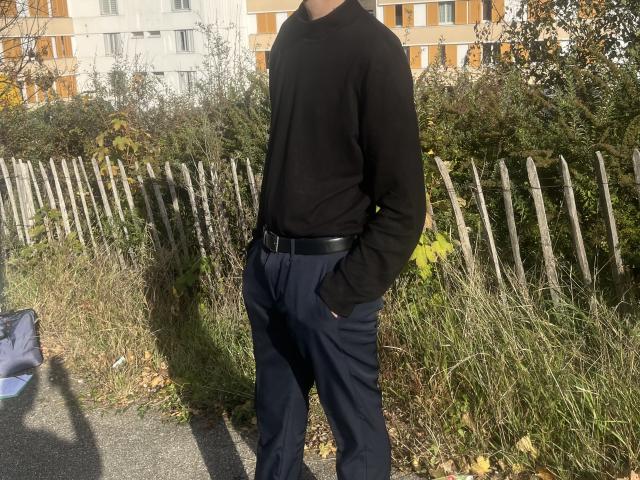 Whitos69 standing in a grassy area, wearing a black sweater and dark pants with hands in pockets, black belt, and a wooden fence in the background.