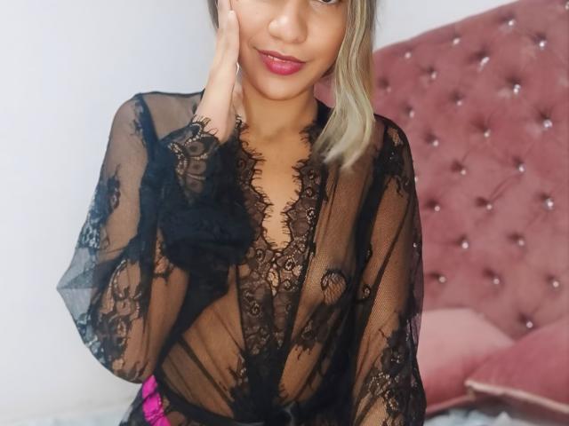 SashaChloe with light brown skin and blonde hair, wearing a sheer black lace robe, showing her tits, hand on cheek, pink lipstick, sitting on pink tufted bed.