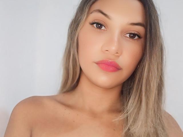 SashaChloe has light brown skin, long blonde hair, brown eyes, and pink lipstick. Her bare shoulders are visible, and she's looking directly at the camera.