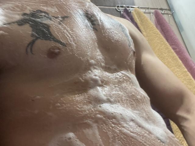 NikkyBanny's wet, soapy chest with visible tattoo above the right nipple, showing a muscular, glistening torso. Her small, erect nipples are partially covered in foam.