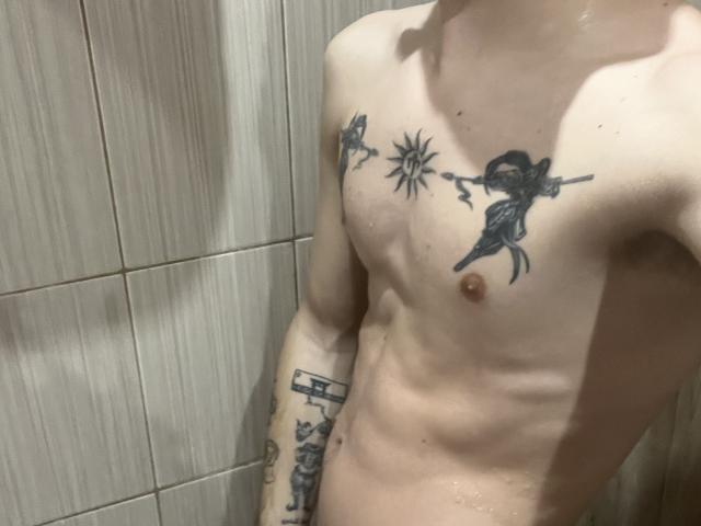 NikkyBanny's shirtless torso with tattoos of a sun, a gun-wielding figure, and a building on their chest and arm, showing a slim, wet, pale body.