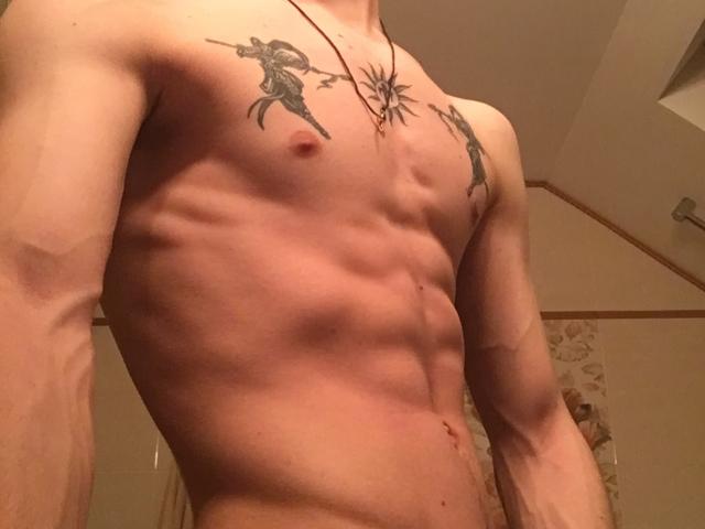 NikkyBanny's skinny, muscular torso with visible ribs and small nipples. Two tattoos of standing figures on chest. Necklace with sun design. Small scar on right side.