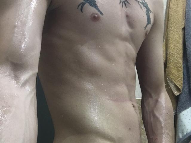 NikkyBanny's wet, muscular torso with visible ribs and tattoos on the chest, glistening skin, and a small, erect nipple. Arms and lower abdomen are in the frame.