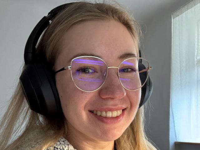 MiaFlirt with blonde hair, wearing big black headphones and gold-framed glasses, has a cute smile and purple light reflections in her eyes. She's looking at the camera.