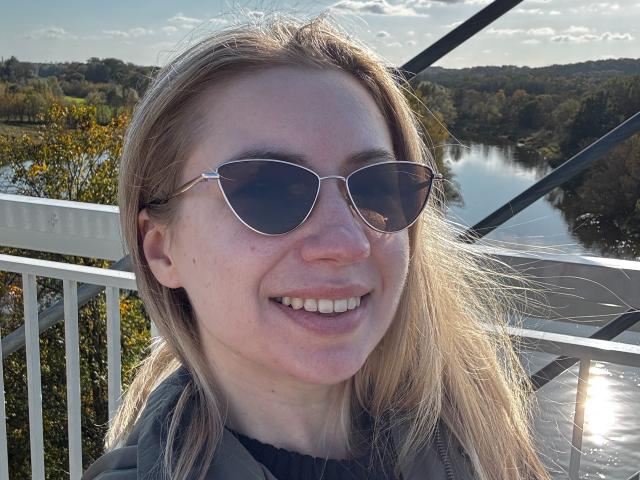 A MiaFlirt tem cabelo loiro, usa óculos de aviador e sorri mostrando dentes brancos. Ela está vestindo um casaco cinza, em um balcão ensolarado, com um rio e árvores ao fundo..
