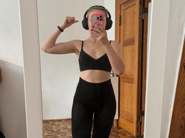 MiaFlirt, vestindo um top de ginástica preto e calças pretas de cintura alta, tem seios pequenos e está tirando uma selfie no espelho com um celular rosa, usando fones de ouvido pretos e apontando para si mesma..